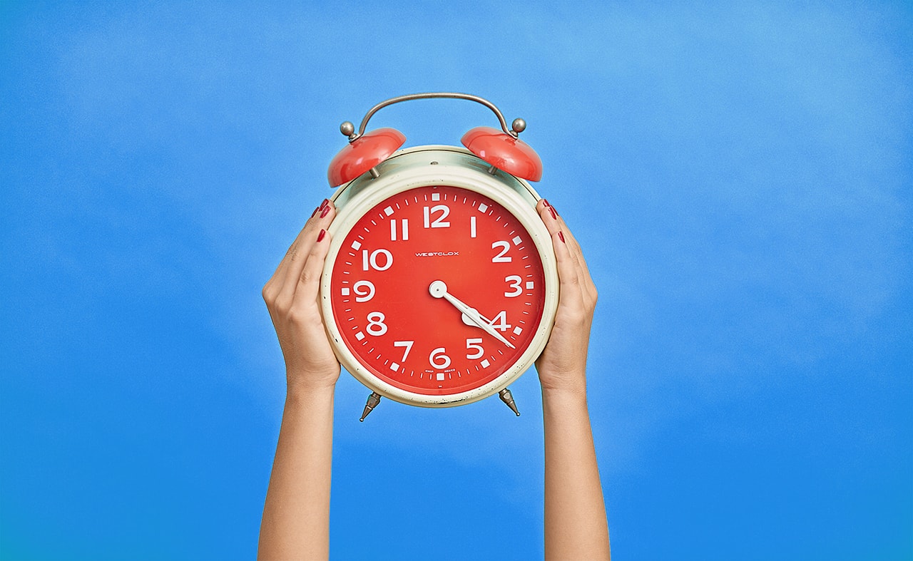 Hands of a woman holding a huge red alarm clock in front of a blue background